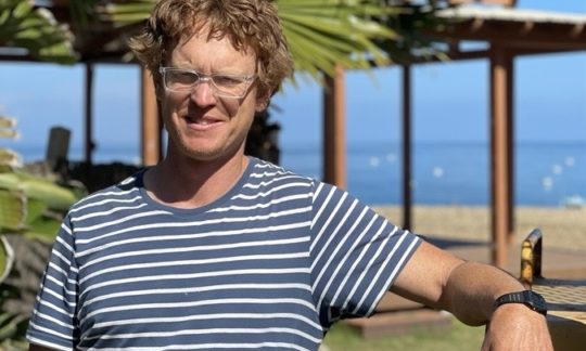 A person wearing glasses and a striped shirt stands outdoors by a palm tree and a building near the beach, evoking the laid-back vibe of summer camp on Catalina Island.