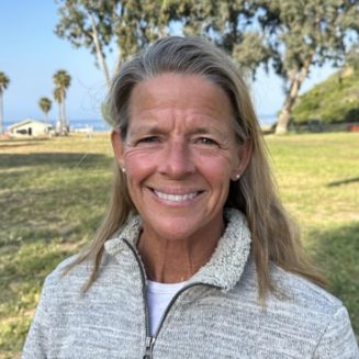 A person with long hair and a white jacket smiles outdoors amidst the lush greenery of a summer camp, with trees and a grassy field in the background.